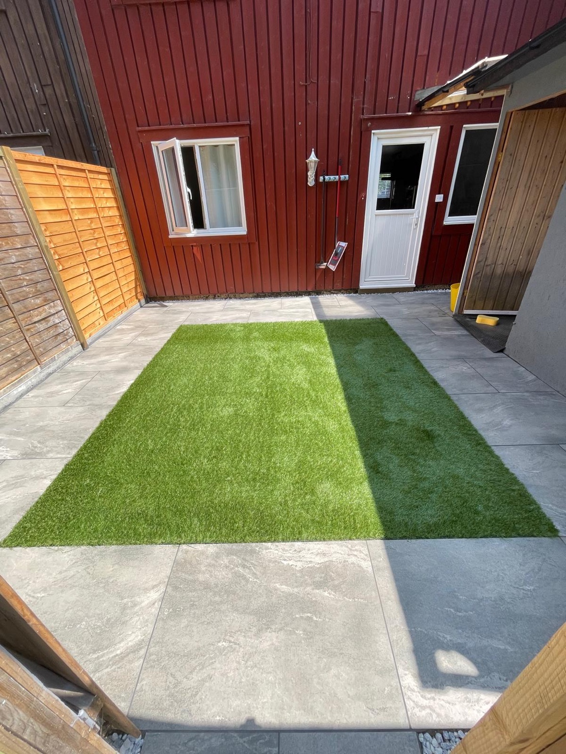 Artificial grass bordered by pale paving in front of a red-clad house